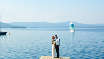 A couple of newlyweds stand on a wooden jetty beside a lake, with sailboats in the background.