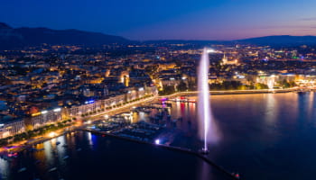 Geneva’s illuminated waterfront and Jet d’Eau seen from above at dusk.